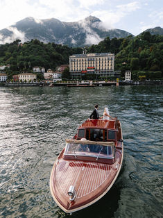 two people are riding on a boat in the water near a large building and mountains