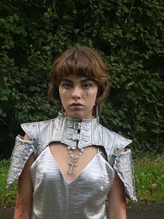 a woman in a silver dress with chains on her neck and shoulder, standing next to some bushes