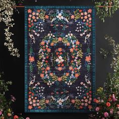 an embroidered wall hanging with flowers and birds in the center, surrounded by greenery