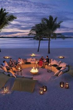 people are sitting around a fire pit on the beach at dusk with palm trees in the background