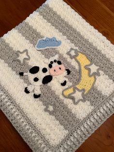 a crocheted baby blanket with a cow and clouds on the top, sitting on a wooden floor