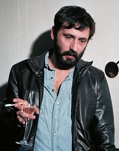 a man holding a wine glass in his right hand and looking at the camera while wearing a leather jacket