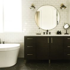 a white tub sitting next to a bathroom sink under a round mirror on top of a wooden cabinet