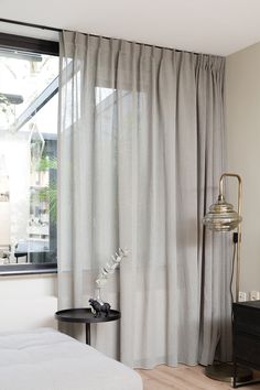 a bedroom with white curtains and a black table in front of the window that has a lamp on it