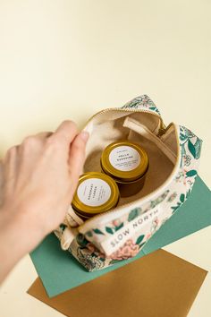 a hand is opening up a small bag with two jars inside it on top of a table