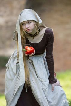 a woman dressed in medieval clothing holding a red rose and looking at the camera while standing outside