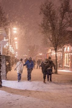 people walking in the snow at night on a city street with buildings and lights behind them