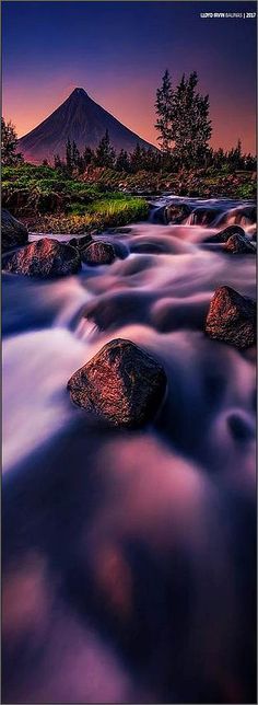 a river running through a lush green forest next to a tall mountain covered in a purple sky