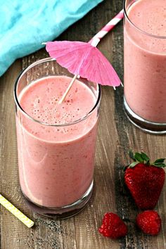 two glasses filled with pink smoothie next to strawberries and an umbrella on a wooden table