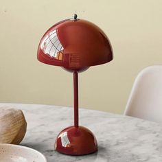 a red table lamp sitting on top of a white table next to a bowl and spoon