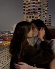 two people kissing each other in front of a cityscape with buildings and lights behind them