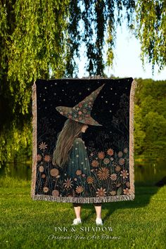 a woman standing in the grass with a witches hat on her head and holding up a tapestry