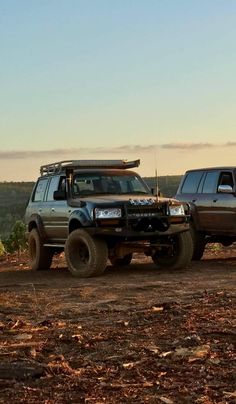 two trucks parked in the dirt near each other on a hill with trees and bushes behind them