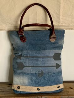 a denim tote bag with arrows on the front and bottom, sitting on a wooden shelf