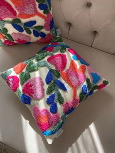 two colorful pillows sitting on top of a white couch next to each other in front of a window