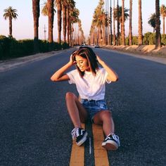 a woman sitting on the side of an empty road with palm trees in the background