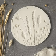 a white plate with dried flowers on it next to some stalks of grass and a rock