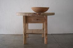 an old wooden table with a bowl on it's top sitting in front of a white wall