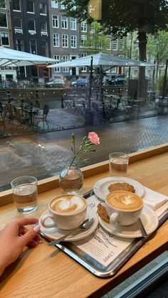 two cups of cappuccino sit on a tray in front of a window