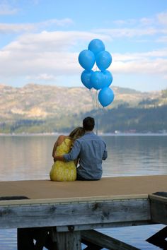 a man and woman sitting on a dock with blue balloons in front of the water