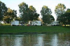 several recreational vehicles parked on the side of a lake in front of some trees and grass