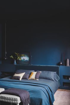 a bed with blue sheets and pillows in a dark colored room, next to a painting on the wall