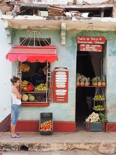 a woman standing in front of a fruit stand on the side of a road near a building