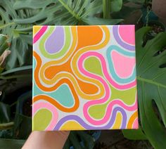 a hand holding up a colorful square painting in front of some plants and leaves on the ground