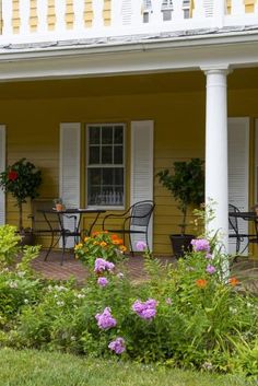 a yellow house with flowers and chairs on the front porch, next to a flower garden