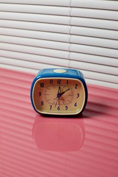 a small blue and yellow clock sitting on top of a pink table next to blinds