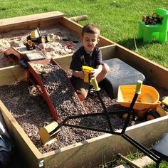 a young boy sitting in a sandbox filled with dirt and gravel holding a drill