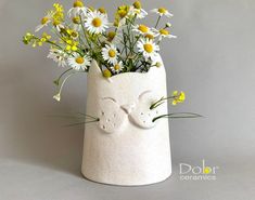 a white vase with flowers in it on a gray surface and the face of a cat made out of clay