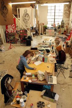 two women sitting at a table in an art studio working on some paper and paint