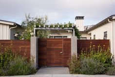 a wooden gate in front of a house with plants growing on the fence and bushes around it