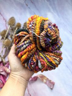 a hand is holding a colorful piece of yarn in their left hand, with dried flowers in the background