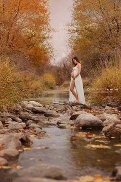 a pregnant woman standing in the middle of a stream surrounded by rocks and autumn leaves