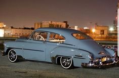 an old blue car parked in front of a tall building at night with the lights on