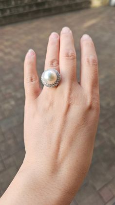 This large white natural freshwater pearl ring is both classic and luxurious in style. The ring setting is made of thick 925 silver, and the ring band is adjustable to fit most wearers. The pearl is a carefully selected brilliant white freshwater pearl with a diameter of 14 to 15 millimeters making it a rare large-sized pearl among freshwater pearls.It has a high-gloss surface with no visible flaws  and rich overtones.  The pearl is surrounded by sparkling small cubic zirconia stones, which furt Fine Jewelry White Pearl Ring Gift, White Oval Pearl Ring As A Gift, White Pearl Ring As Gift, White Pearl Ring Gift, White Oval Pearl Ring Gift, Pearl Drop Diamond Ring, White Gold Pearl Ring As Gift, White Akoya Pearl Round Ring, White Akoya Pearl Ring