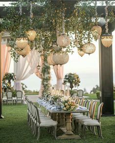 an outdoor dining table with hanging lights and flowers on the top is surrounded by greenery