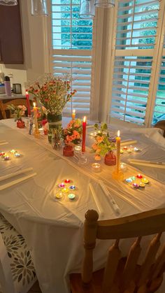 a table with candles and flowers on it in front of the window is set for dinner