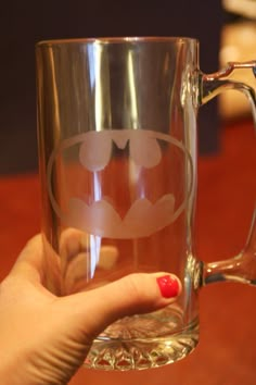 a person holding up a glass mug with a batman symbol on the side and red nail polish