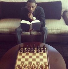 a man sitting on a couch reading a book next to a chess board and table