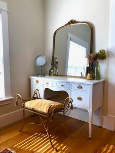 a white dresser with a mirror, chair and vase on it in a room next to a window