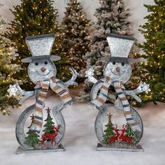 two metal snowmen with hats and scarfs standing in front of christmas tree decorations