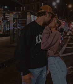 a man and woman standing next to each other on a street at night with buildings in the background