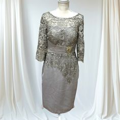 This Elegant Dress Features A Fitted Silhouette In A Soft Taupe-Gray Hue. The Top And Sleeves Are Adorned With Intricate Lace Detailing, Adding A Touch Of Sophistication. The Three-Quarter Sleeves Offer A Classic Look, While The Waist Is Accentuated By A Satin-Like Band, Gathered At The Center With A Jeweled Brooch, Giving The Dress A Refined And Polished Finish. Perfect For Formal Events Or A Special Occasion Where Understated Elegance Is Key. Silver Color With Sage Green Hues. 28 Inches Waist Gray Knee-length Wedding Dress, Fitted Gray Dress For Mother Of The Bride, Elegant Gray Dress For Mother Of The Bride, Elegant Gray Mother Of The Bride Dress, Elegant Knee-length Gray Dress, Elegant Gray Knee-length Dress, Gray Knee-length Formal Dress, Jeweled Brooch, Formal Dress For Women