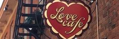 a large red heart shaped sign hanging from the side of a brick building that says love cafe