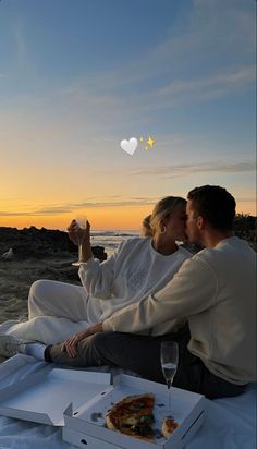 a man and woman sitting on the beach drinking wine, eating pizza and watching the sun set