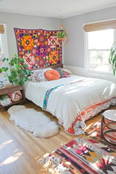 a bedroom with a bed, rugs and plants on the floor in front of two windows