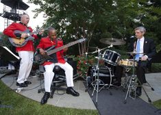 two men in red jackets and white pants are playing instruments while another man is sitting on a chair